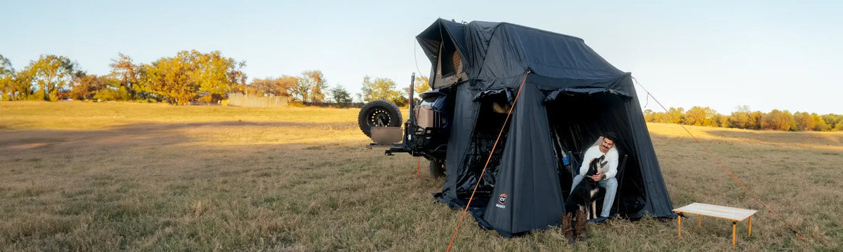 A person sits with a dog at the entrance of a rooftop tent attached to a vehicle, set up in a grassy field with trees and a bench nearby under a clear sky.