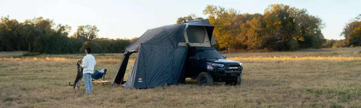 A person stands by a camping chair and dog near a black truck with a rooftop tent set up in a grassy field bordered by trees.