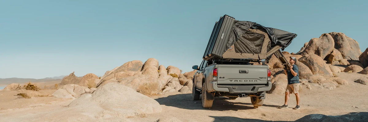 A person stands next to a gray Toyota Tacoma with a rooftop tent set up in a rocky desert landscape under a clear blue sky.