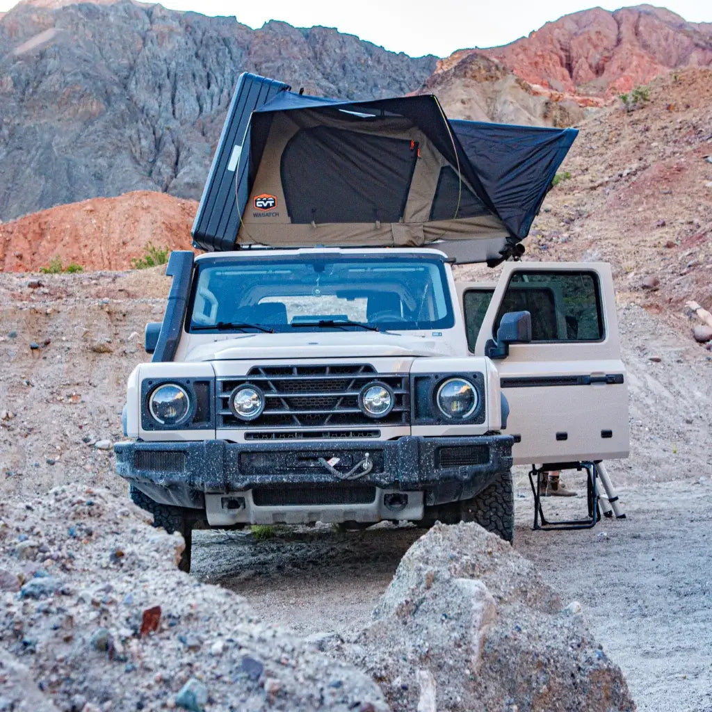 A white off-road vehicle with a rooftop tent is parked in a rocky desert area, with one door open and rugged mountains in the background.